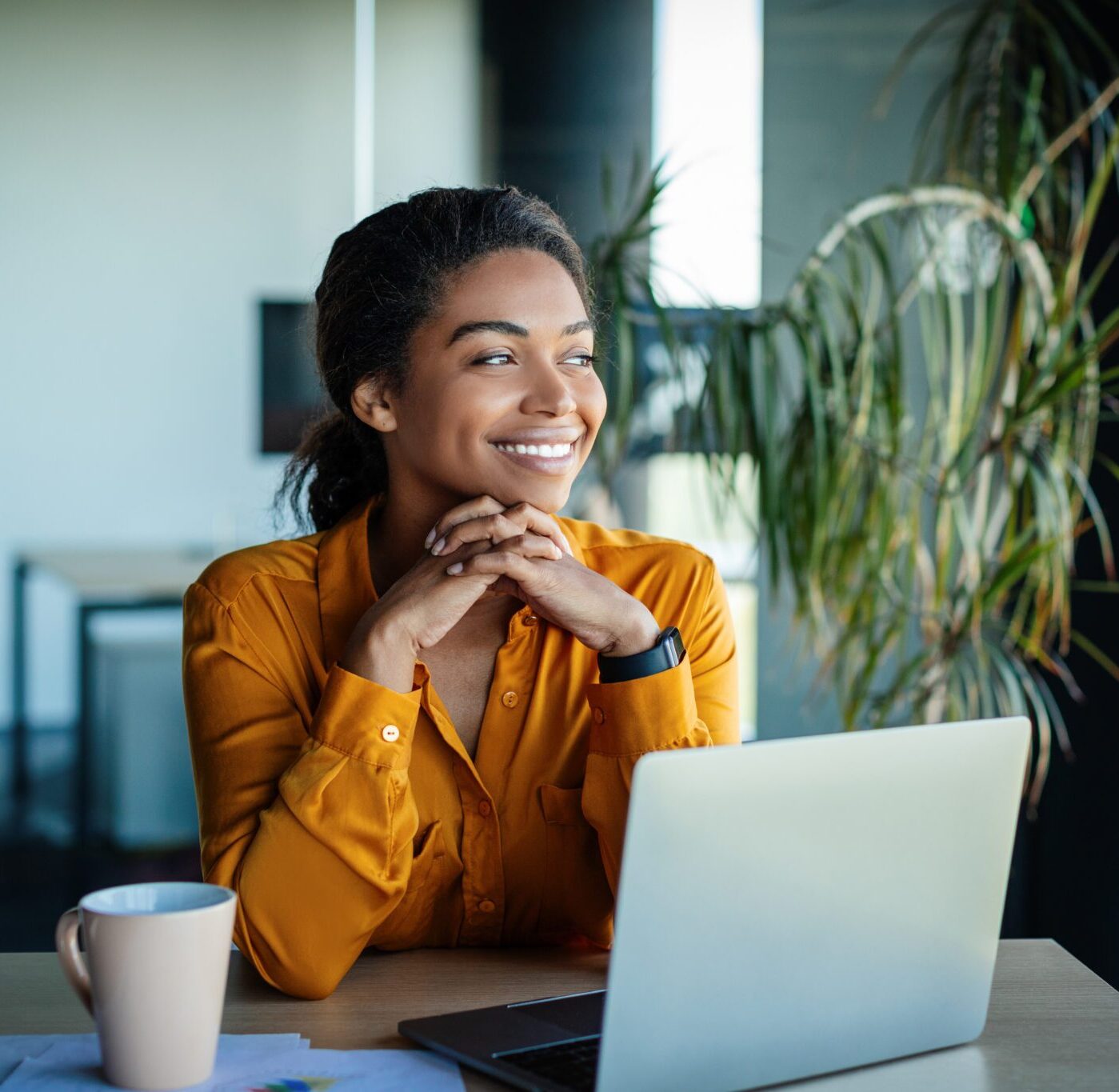 Portrait of dreamy african american businesswoman sitting at desk in office and thinking while working on laptop, looking away with pleased face expression, copy space
