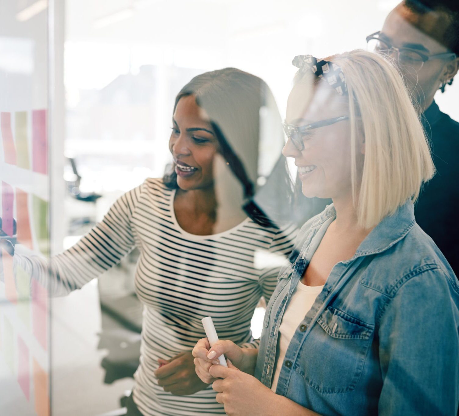 Group of diverse young work colleagues smiling while brainstorming together with sticky notes on the glass wall of a modern office